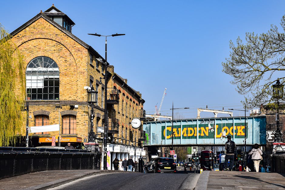 The image shows a busy street scene in Chiswick, with a prominent brick building featuring large arched windows on the left side, and a bridge with a blue metal structure marked 'CALDEIN LOCK' spanning across the road. Several pedestrians are walking along the pavement, and parked cars line the street. In the foreground, a flatbed trolley with black straps is being used to carry moving boxes wrapped in plastic and packing materials, suggesting a home relocation or furniture transport process. The loading area is part of an indoor space, possibly a property entrance, with some boxes and furniture partially visible inside or near the doorway. The scene is set during daylight with clear skies, and trees with budding leaves are visible on the right side, indicating a springtime relocation effort. Man with Van Chiswick is engaged in the packing and loading process, utilizing equipment like trolleys and blankets for moving furniture and boxes efficiently.