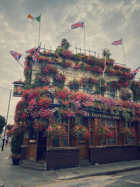 A multi-storey pub building decorated extensively with vibrant pink, red, and purple flowering plants covering the facade, balconies, and windows. The building has several Union Jack and an Irish flag flags flying from flagpoles on the roof, and a sign identifying it as the Churchill Arms. The ground floor features wooden-framed windows and doors, with potted plants and flowers placed near the entrance. Inside the pub, visible through the windows, is the interior lighting with a warm glow. Outside, the surrounding pavement is visible with some pedestrians and street furniture. The scene is captured during daylight with a cloudy sky overhead. The pub is situated in a busy street, possibly part of a home relocation or furniture transport process, with arrangements indicative of a well-known local establishment in Chiswick.