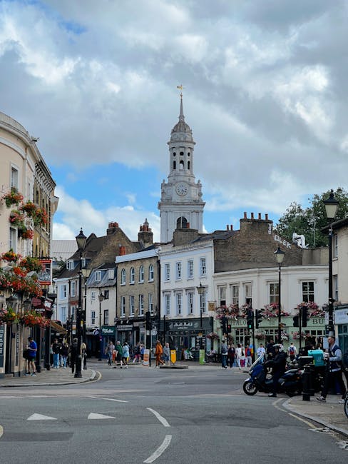 The image shows a busy street scene in Chiswick, with a prominent brick building featuring large arched windows on the left side, and a bridge with a blue metal structure marked 'CALDEIN LOCK' spanning across the road. Several pedestrians are walking along the pavement, and parked cars line the street. In the foreground, a flatbed trolley with black straps is being used to carry moving boxes wrapped in plastic and packing materials, suggesting a home relocation or furniture transport process. The loading area is part of an indoor space, possibly a property entrance, with some boxes and furniture partially visible inside or near the doorway. The scene is set during daylight with clear skies, and trees with budding leaves are visible on the right side, indicating a springtime relocation effort. Man with Van Chiswick is engaged in the packing and loading process, utilizing equipment like trolleys and blankets for moving furniture and boxes efficiently.