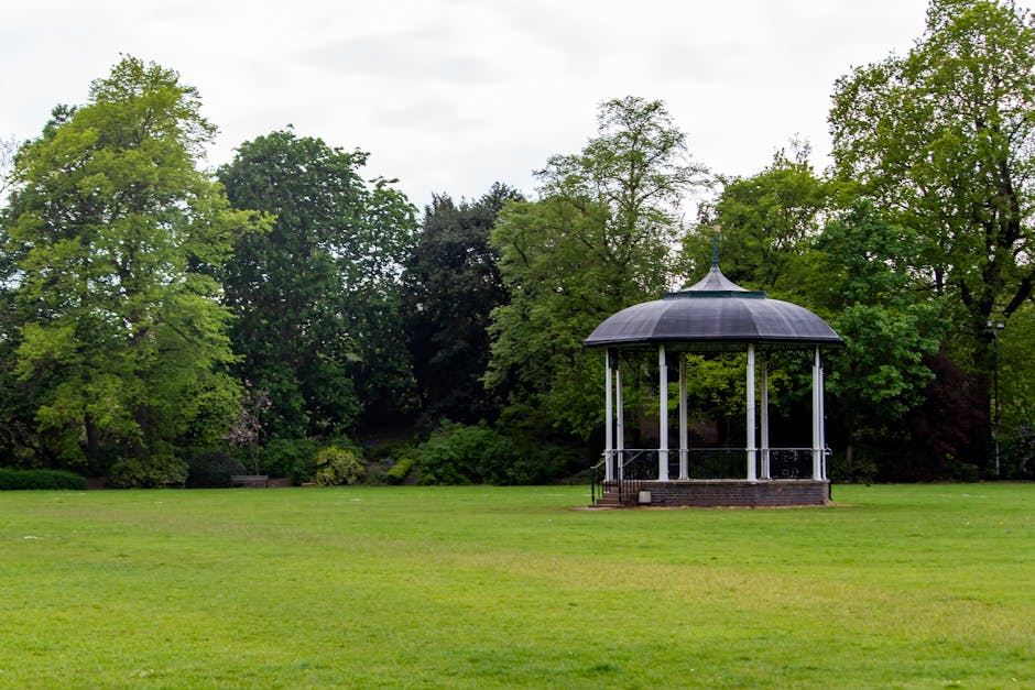 A small, circular bandstand with a dark, domed roof supported by white columns, situated on a brick platform within a park setting. The surrounding area features a well-maintained grassy lawn, bordered by a variety of mature trees with green foliage, some with lighter shades and others darker, indicating different species and growth stages. The park is outdoors under an overcast sky, providing diffuse natural light. In the background, dense tree cover creates a natural backdrop, and the open space suggests a peaceful environment suitable for outdoor gatherings or leisure activities. This scene is relevant to home relocation processes, with the bandstand seen from a distance as part of the scenic environment that may be accessed during a house move or property viewing, which is supported by Man with Van Chiswick’s house removals services. No vehicles or moving equipment are visible in this image, only the natural park setting and the bandstand itself.